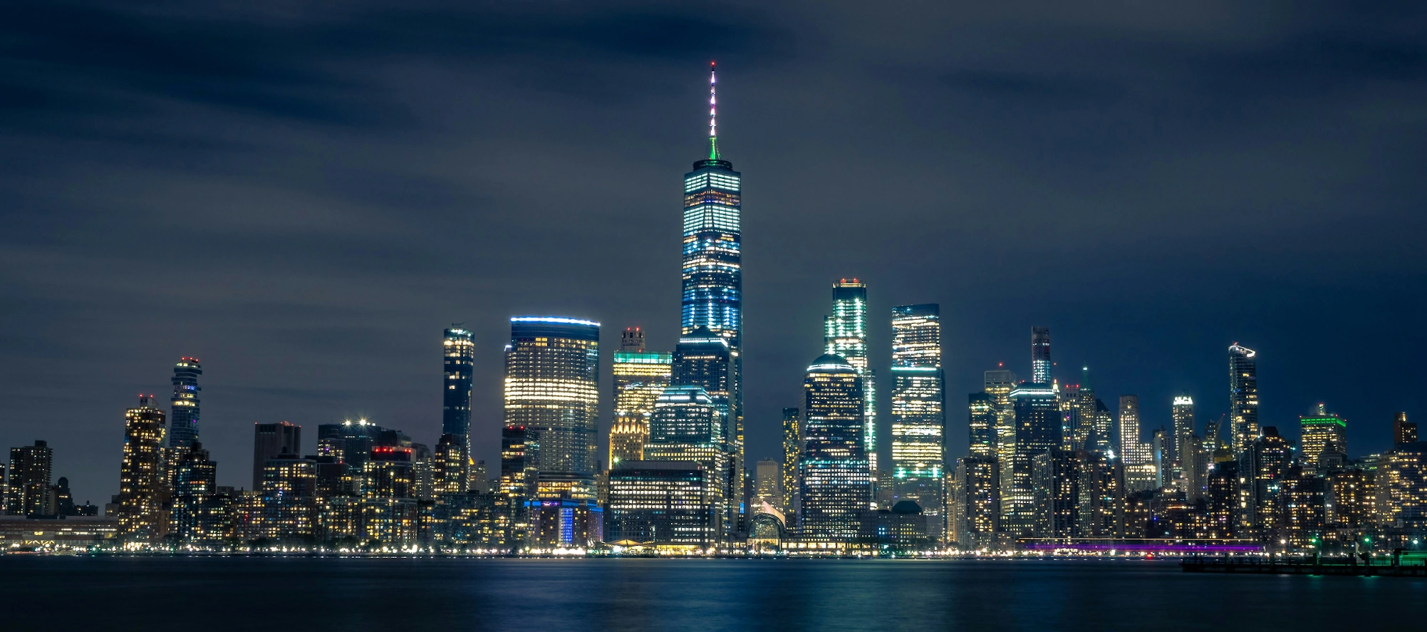 Night view of the New York City skyline with illuminated buildings and reflections on the water.