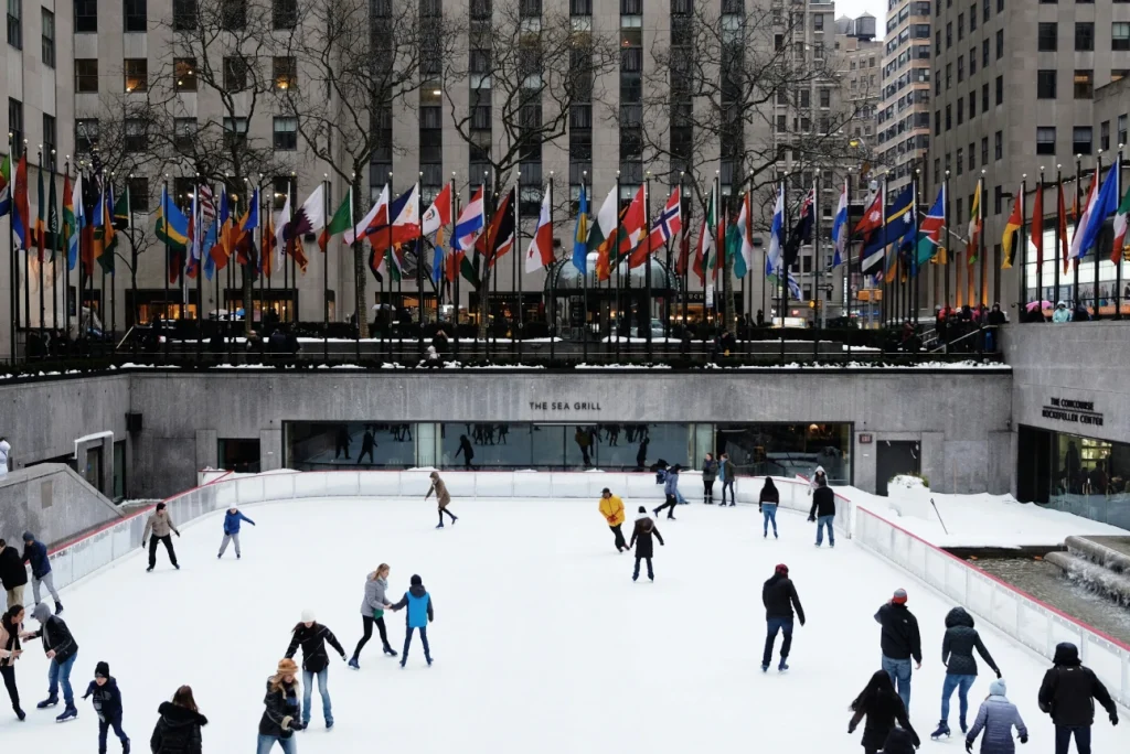 People ice skating on The Rink at Rockefeller Center, a famous winter attraction in NYC, surrounded by towering skyscrapers and festive lights.